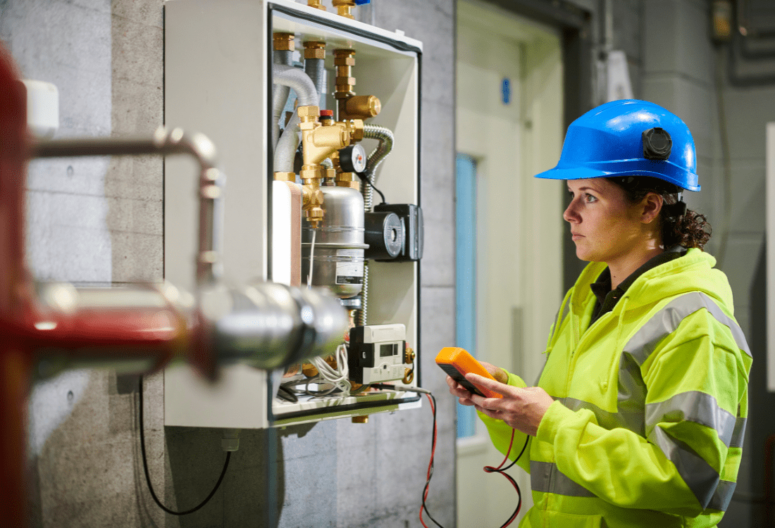 A recently certified electrician, a young woman clad in safety gear, is fully immersed in meticulously and attentively recording readings from an electrical system.